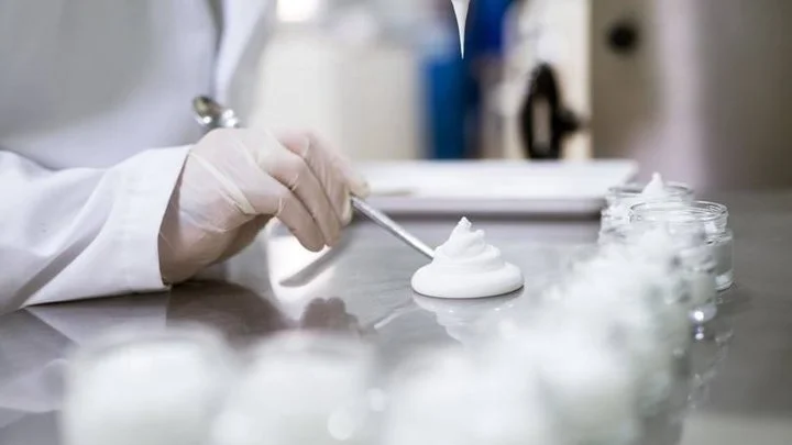 Lab technician wearing gloves examining cream texture on a metal table with jars of cream in the background.